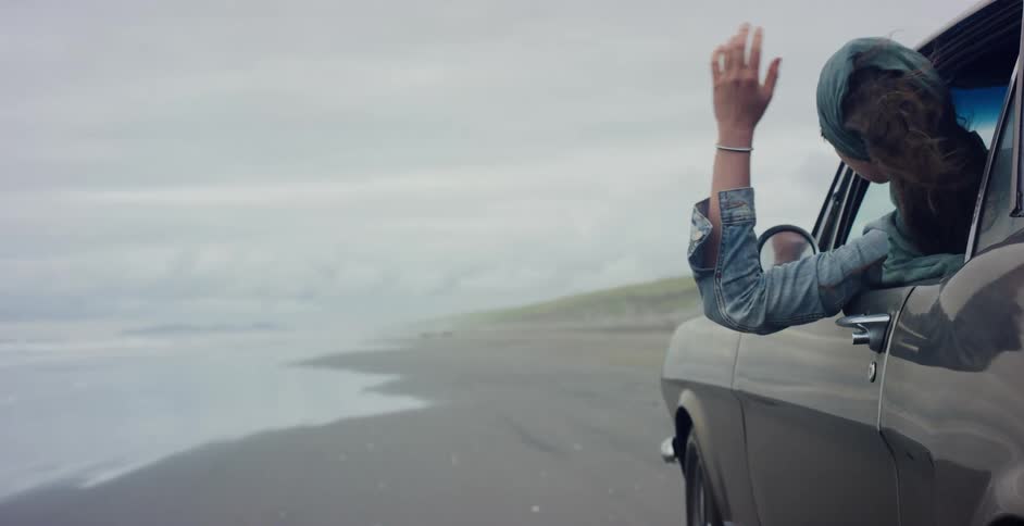 A woman with a headscarf enjoys the coastal breeze as she leans out of a classic car driving along a sandy shore in slow motion