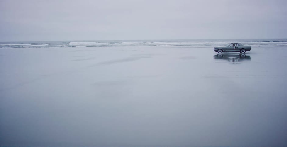 Wide shot of a vintage American car parked on a serene, misty beach, reflecting on the wet sand