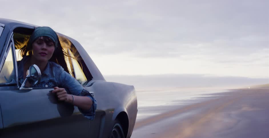 A young woman enjoys the ride as she leans out the window of a classic car driving along the beach at dusk