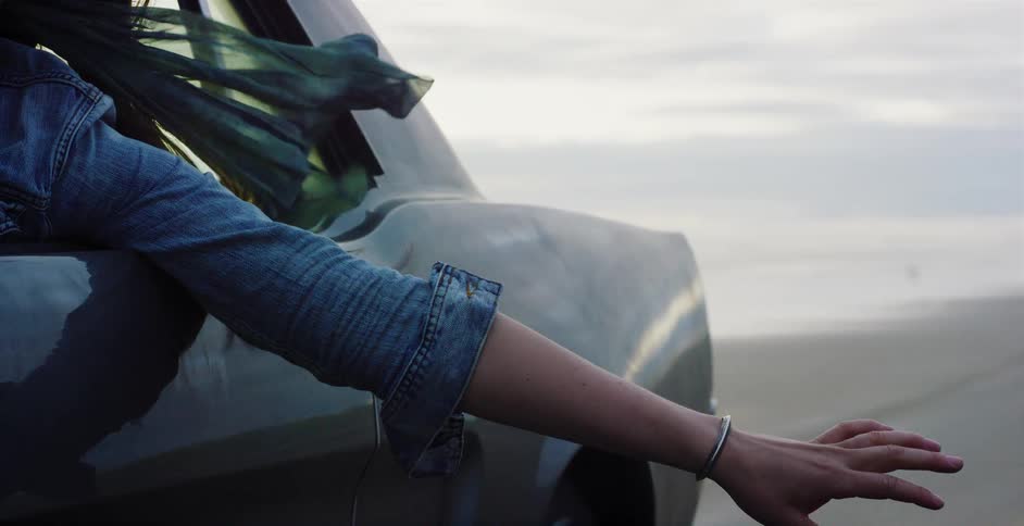 Slow-motion close-up of a woman's arm out the window of a classic car as her scarf blows in the wind on a coastal road trip