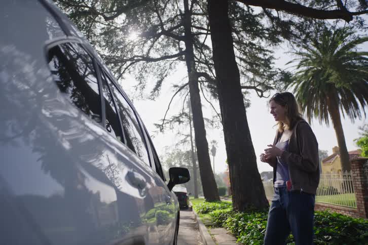 A woman standing on a sunny, tree-lined suburban sidewalk has a conversation with someone inside a parked car