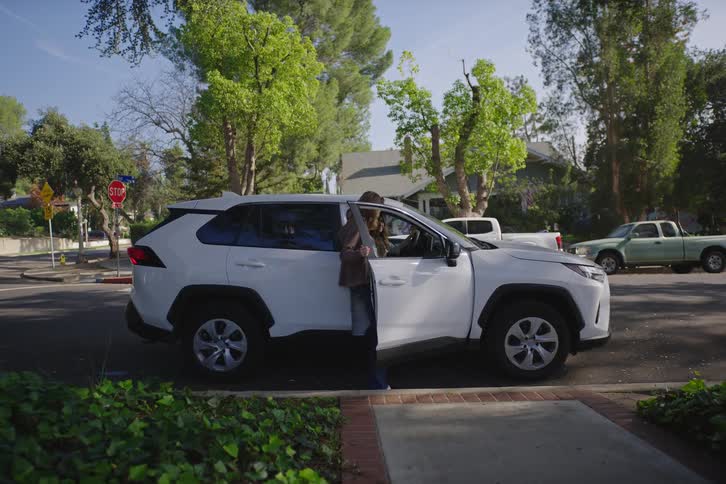 A long-haired person walks to a parked white SUV on a suburban street and gets into the passenger seat on a sunny day