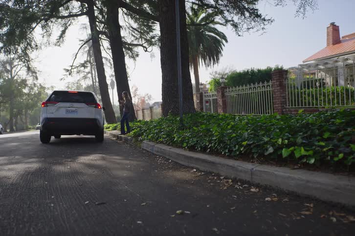 A woman waits by her parked SUV on a quiet, tree-lined suburban street on a sunny day