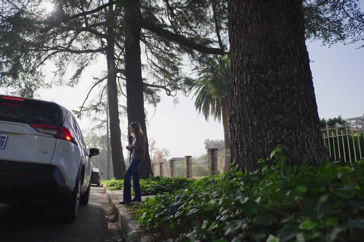 A woman waits on the curb next to her parked SUV on a sunny, tree-lined street while using her smartphone