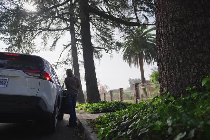 A woman walks down a tree-lined sidewalk and gets into the driver's seat of her parked white SUV