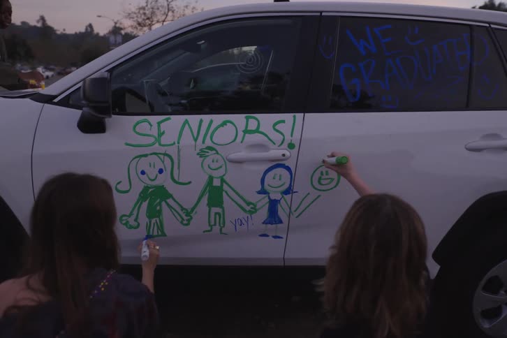 A diverse group of graduating high school seniors celebrate by drawing festive messages and pictures on a white car at dusk
