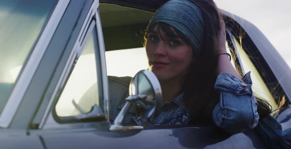 A beautiful young woman in a denim jacket and headband smiles softly from the driver's window of her classic car at sunset