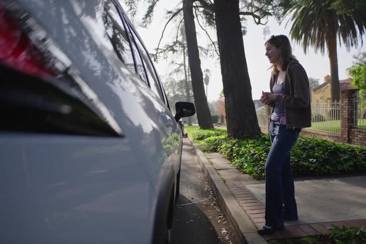 A woman stands on a sunny suburban sidewalk and has a pleasant conversation with someone inside a parked car