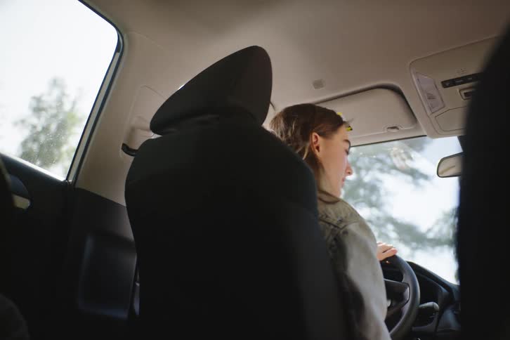 Backseat view of a happy young woman driving and chatting with a passenger during a sunny car ride