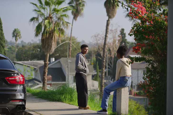 A teenage boy and girl stand on a sidewalk and have a conversation in a quiet suburban neighborhood with palm trees