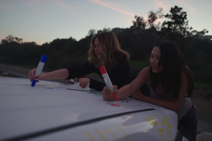 Slow motion of happy young women drawing on the roof of their van with colorful markers at twilight
