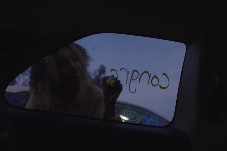 A young woman seen from inside a vehicle happily writes 'Congrats!!' and draws a smiley face on the car window with a yellow marker at dusk