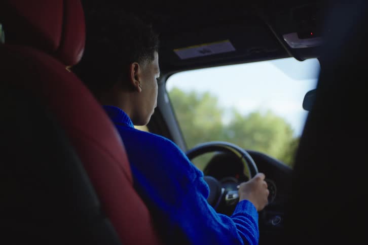 An over-the-shoulder shot of a young Black man driving a modern car with a red interior on a sunny day