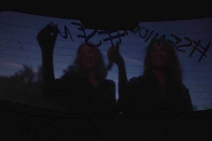 Two high school senior girls laugh while writing celebratory hashtags on their car window at dusk