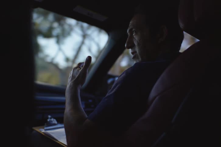Over-the-shoulder view of a man in the passenger seat of a moving car, gesturing with a pen as he gives instructions