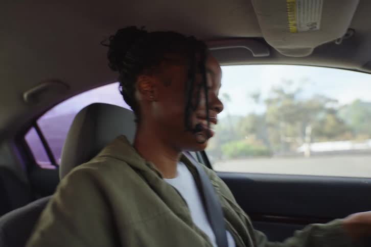 A happy young Black woman gets in the driver's seat of her car, starts the engine, buckles her seatbelt, and enjoys a sunny day drive