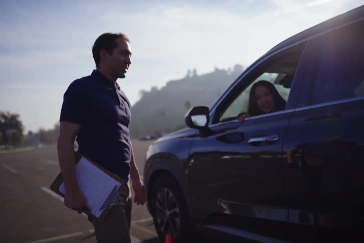 A male driving instructor happily gives directions to a female student as she practices backing up through cones on a sunny day