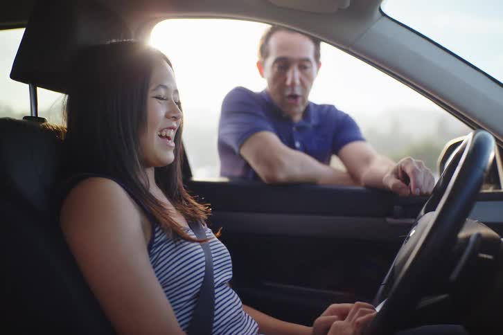 A teenage girl smiles and laughs while her father gives her a driving lesson, before her expression turns more serious as she focuses