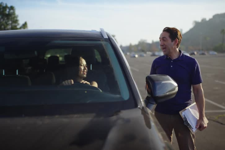 A friendly driving instructor gives a lesson to a young woman learning to drive in a car in a large, empty parking lot