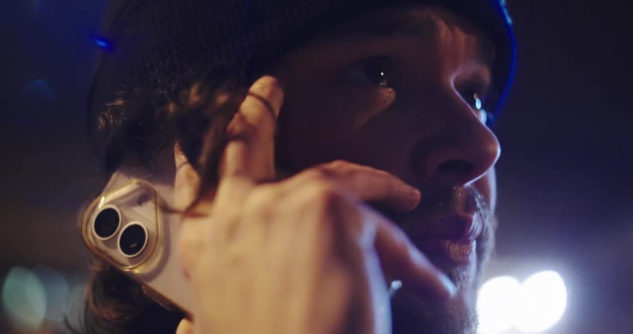 A dramatic slow-motion close-up of a man in a beanie talking on his smartphone with city lights in the background
