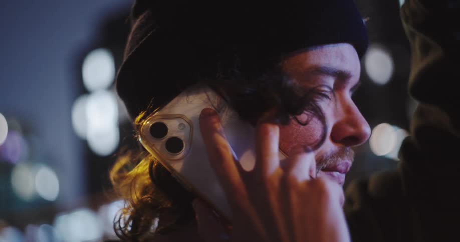 Slow motion close-up of a young man in a beanie talking on his smartphone against the blurred city lights at night