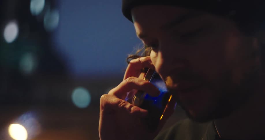 A serious young man in a black beanie has an intimate phone conversation on a city street at night