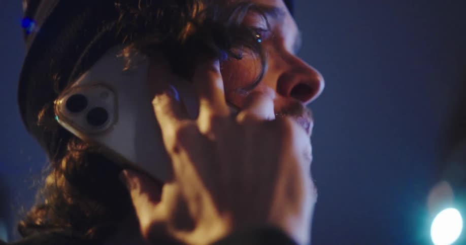 An emotional close-up of a worried young man in a beanie talking on his smartphone at night