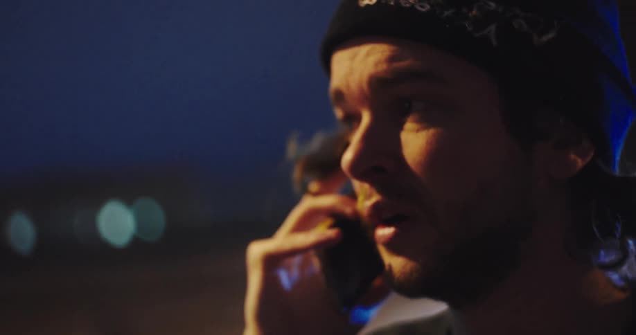A concerned man in a beanie has a serious conversation on his phone at night with city lights blurred in the background