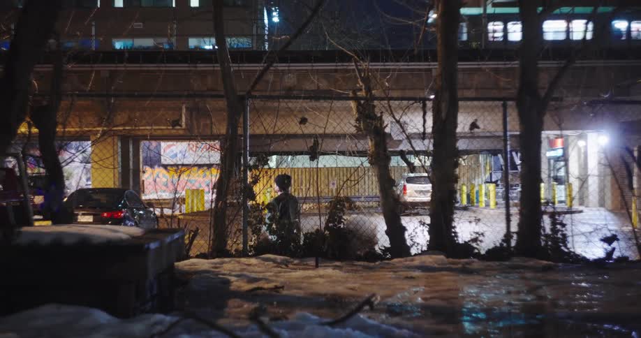 At night, a solitary person forages in a snowy urban lot while a city train passes on the elevated tracks above