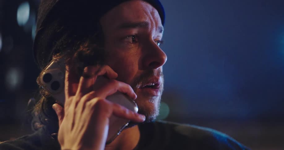 A close up on a distraught man wearing a beanie as he receives bad news over the phone at night