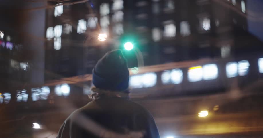 A moody, out-of-focus shot of a pensive young man standing on a city street at night as a train passes by in the background