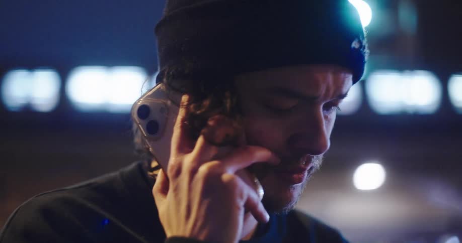 A close-up shot of a young man with a worried expression having a serious phone conversation on a city street at night