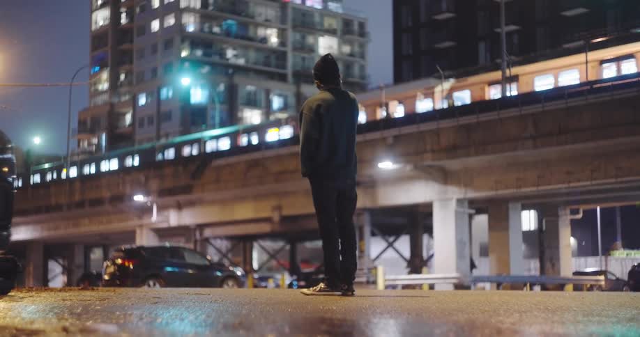 A cinematic shot of a man standing in the city at night, watching a train pass on an overpass before looking at his phone