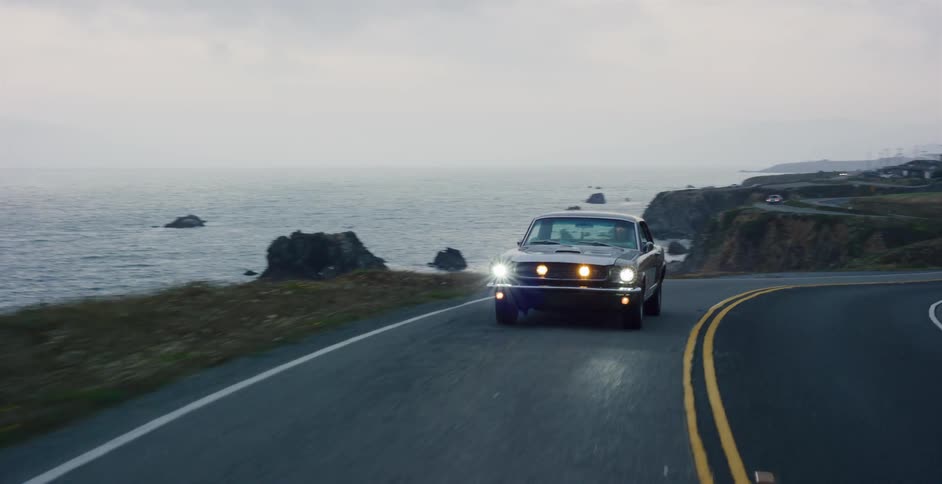 A classic muscle car with headlights on cruises down a winding coastal highway next to the ocean on a foggy evening