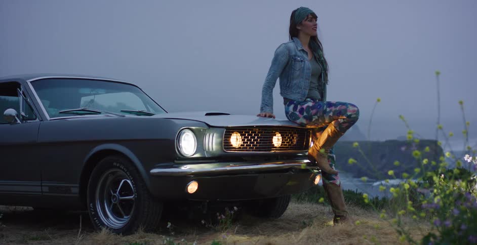 A stylish woman sits on the hood of a classic Ford Mustang parked on a foggy coastal cliff at dusk