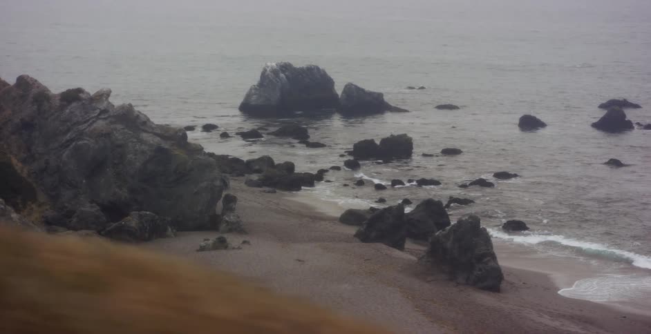 A moody and atmospheric tracking shot along a rugged, rocky coastline shrouded in dense fog
