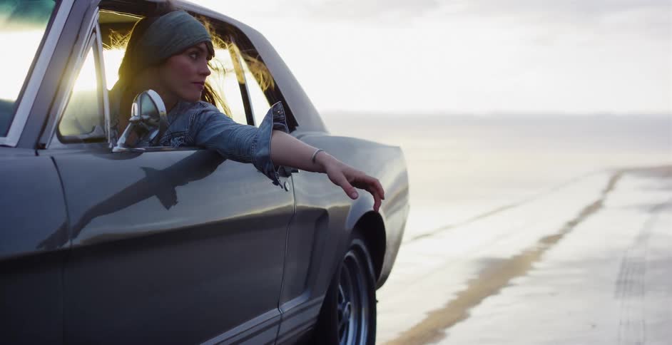 A free-spirited woman with wind in her hair relaxes in a classic car driving along the beach during a hazy sunset