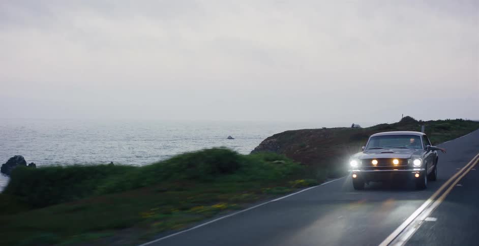 A vintage black car cruises down a scenic coastal highway with its headlights on at dusk