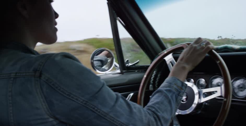 A woman with long hair drives a classic convertible car down a coastal highway, the wind blowing through her hair in slow motion