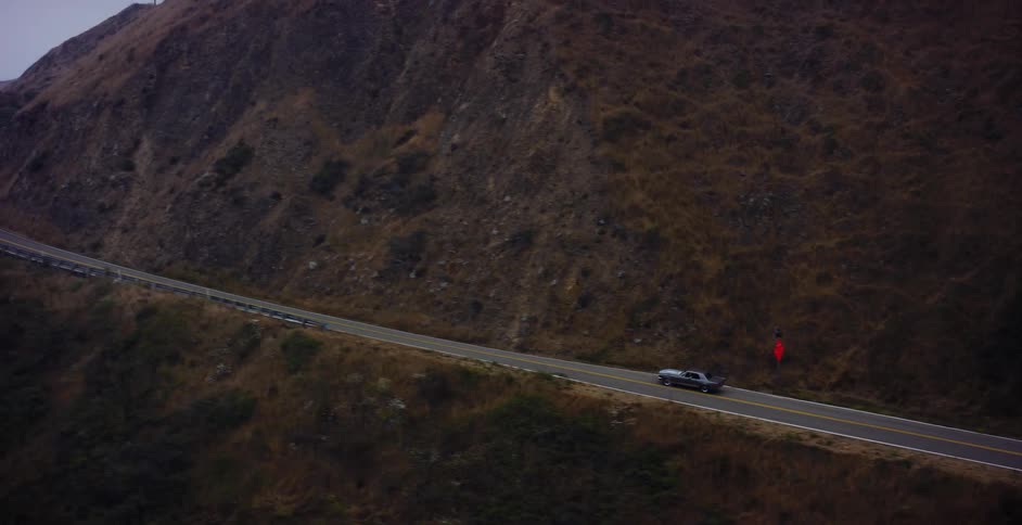 An aerial view tracks a vintage muscle car as it drives along a winding coastal highway through rugged, hilly terrain