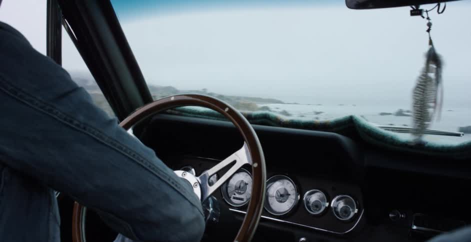 A woman's hands grip the wooden steering wheel of a classic car while driving along a foggy, scenic coastal highway