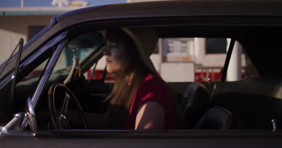 A stylish woman in a wide-brimmed hat parks her classic muscle car at a desert gas station and gets out in slow motion