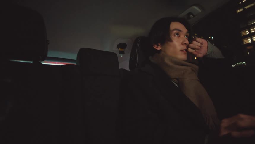 A pensive young man wearing a scarf looks out the window at the city lights while riding in the back of a car at night