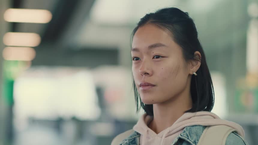 Close-up portrait of a thoughtful young Asian woman standing still in a blurred public transit station