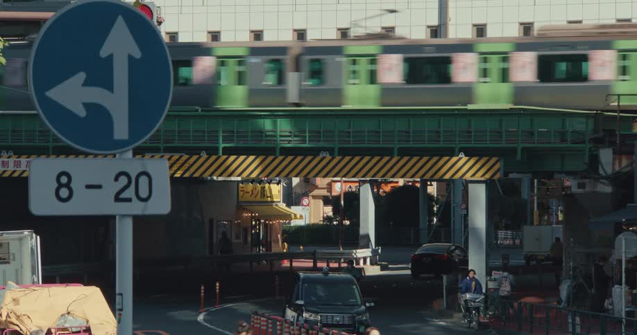 A green commuter train travels along an elevated track over a busy underpass on a city street in Tokyo, Japan