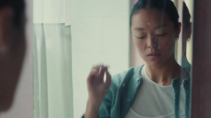 A young Asian woman with acne cream on her face ties her hair back while looking in the mirror