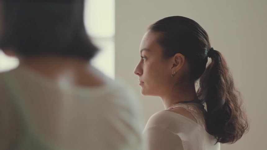 A woman with her hair in a ponytail peacefully meditates with eyes closed in a calm, sunlit studio