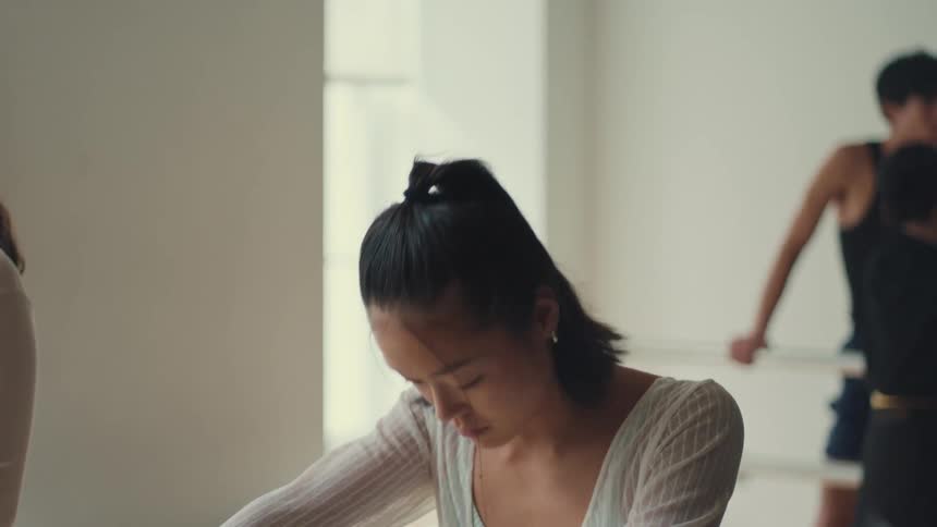 Close-up slow-motion shot of a focused young Asian ballerina during a moment of introspection in a brightly lit dance studio