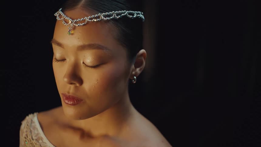 A contemplative Asian ballerina with a bejeweled tiara closes her eyes in a dramatic, softly lit close-up against a black background