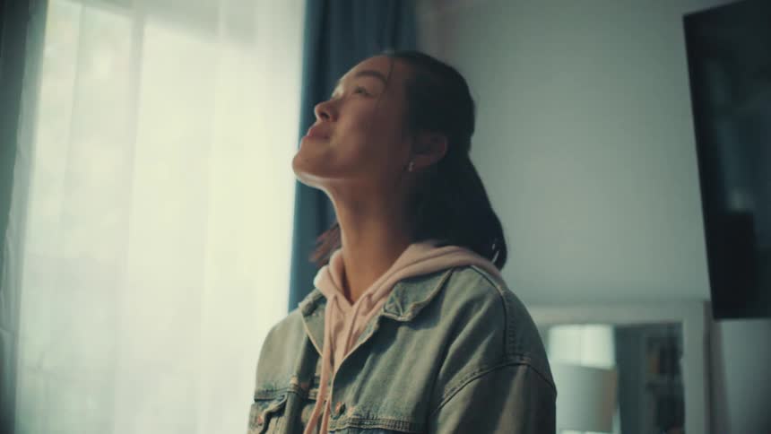 Happy young Asian woman smiles while texting on her smartphone before looking up to take a relaxing breath in her sunlit room